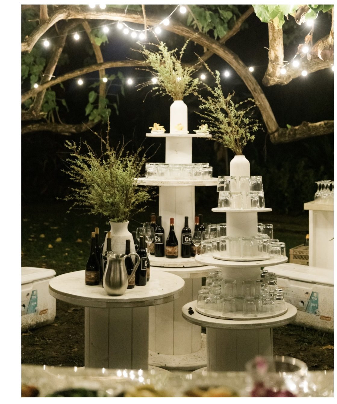 White tiered bar shelving unit with glassware, bottles, and greenery at a nighttime event