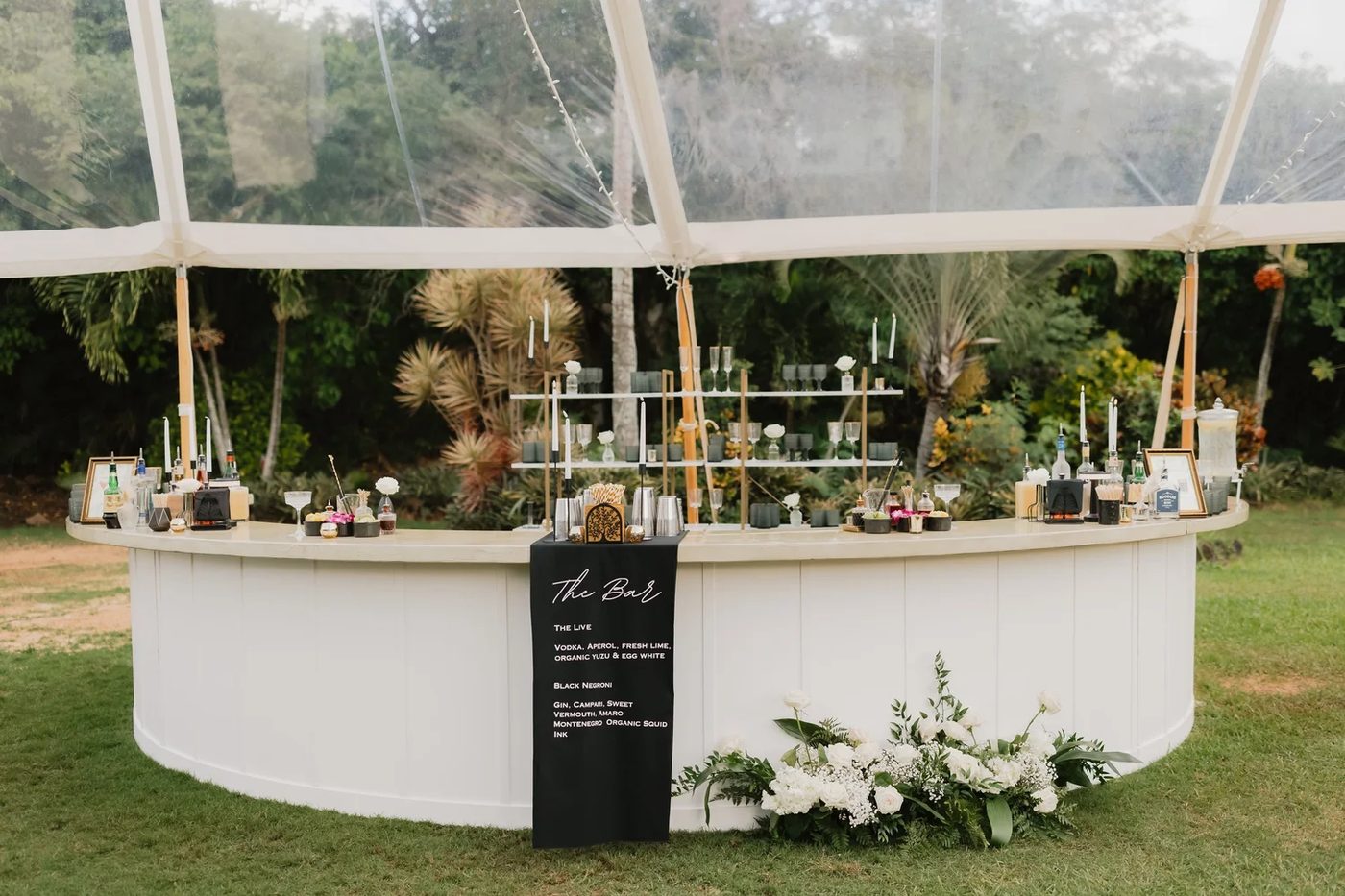 White semi-circle bar fully styled with back bar shelving, cocktail menu, and florals under a clear tent
